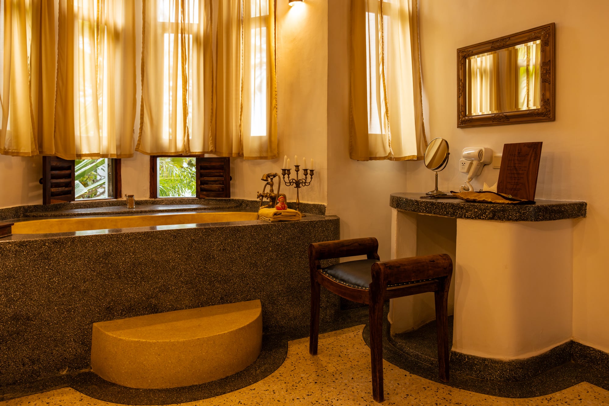 The Jua bathroom: a deep bath set into terrazzo, tall curtained windows, a dressing table and mirror.
